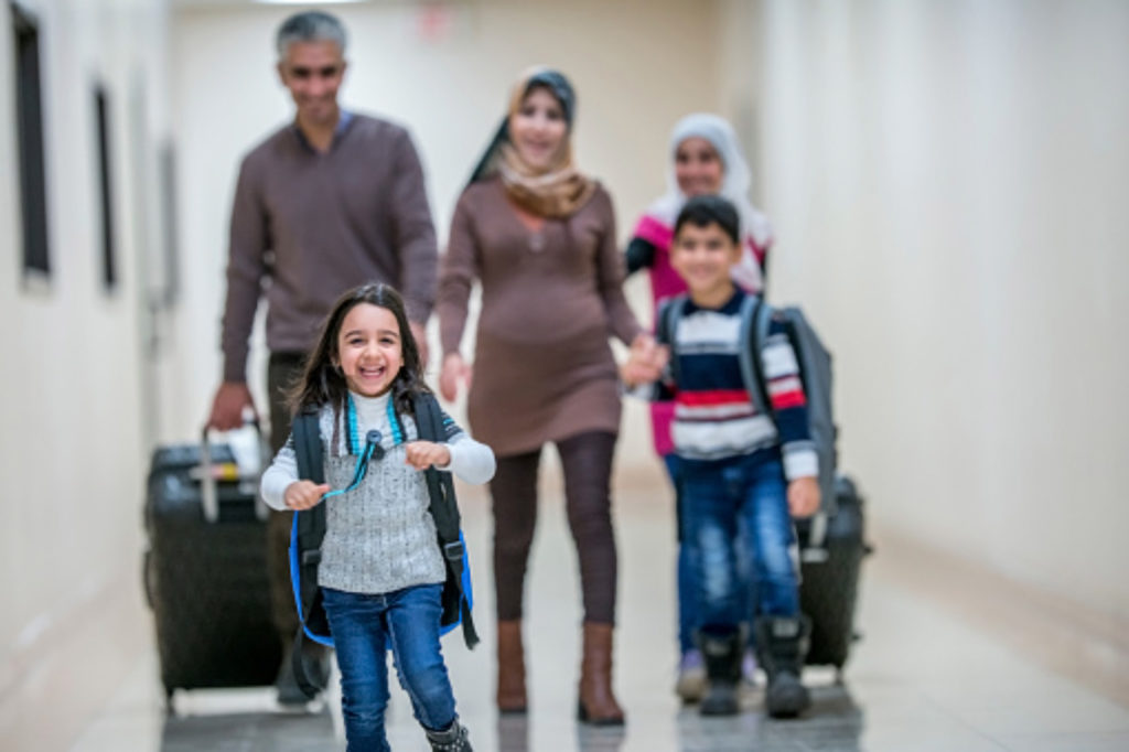 A Middle-eastern father, mother, brother and two sisters have just arrived to their new apartment building. They are carrying their luggage with them. They are walking down the hallway, being lead by the happy youngest daughter.