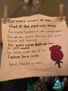 Words written on fabric above a drawing of a rose. Text reads: "For every scream of help/ that in the past was silenced/ For every injustice in our history hidden./ For all the scars that are left and we're still healing./ For every person that right now is still scared./ For every single one of us/ Fighting for a change/ Here, there's a rose.